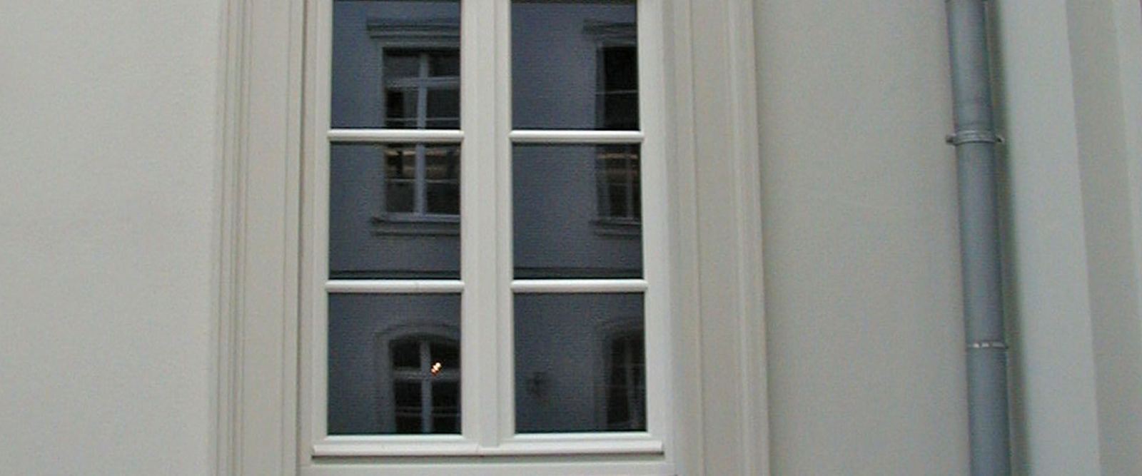 Zwei hohe Sprossenfenster in einer hellen Putzfassade, daneben ein Fallrohr.